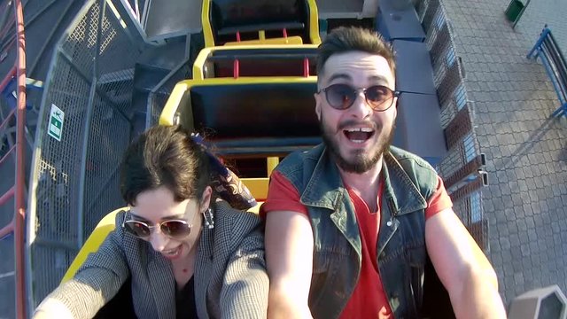 Selfie video of couple riding the roller coaster. Hipster man and stylish woman have fun at amusement park rides. Friends extreme riding, laughing and screaming of excitement, adrenalin.