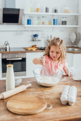 selective focus of child mixing flour in glass bowl with whisk near bottle of milk, rolling pin and eggs on kitchen table