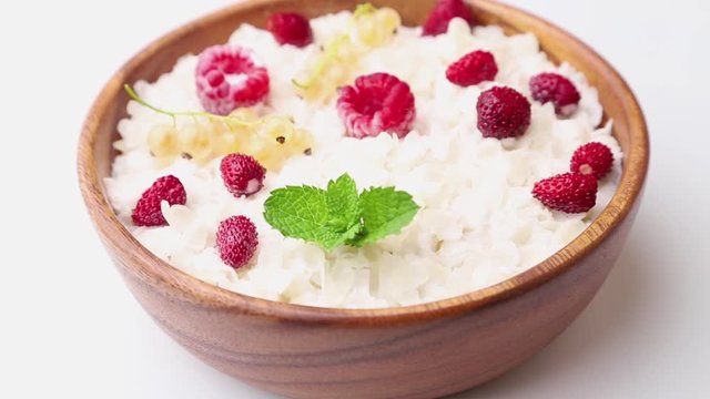 Rotating of rice flakes porridge with milk and strawberry in wooden bowl on white background. Side view, close up.