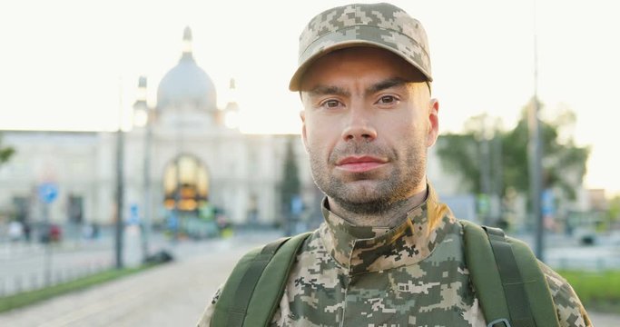 Portrait of serious Caucasian handsome young male soldier in cap with backpack standing at street. Man militarian looking at camera at train station. Bus stop. Military uniform. Zooming in.