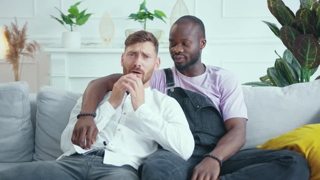 Two diverse gay friends watching drama film on TV at home. Sensitive caucasian man crying with tears and afro-american boyfriend supporting him.