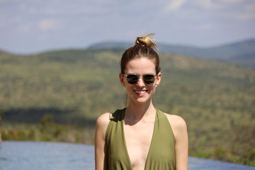 Young woman in swimsuit wearing sunglasses next to infinity pool