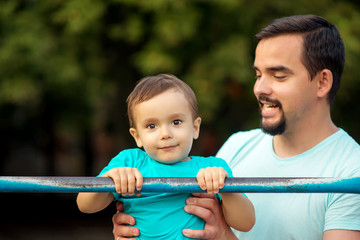 Fototapeta na wymiar Father teaching toddler son to do pull ups exercise on steel bar outdoors. Healthy family, parental support and sports for kids concept. Copy space