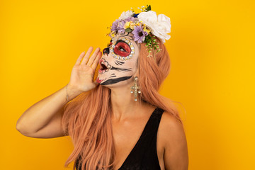 young Caucasian woman sugar skull make up, standing against yellow background profile view, looking happy and excited, shouting and calling to copy space.