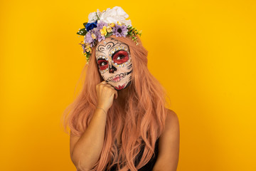 Face expressions and emotions. Thoughtful young pretty girl wearing sugar skull Halloween make up holding hand under her head, having doubtful look.