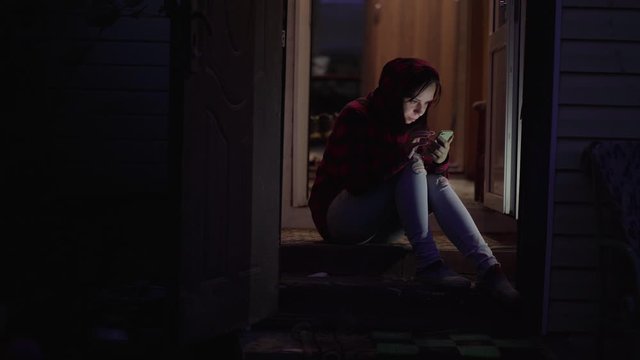 Young woman sitting on porch and browsing mobile phone at night. Adult female flipping chat in smartphone, sitting on porch of house in night.