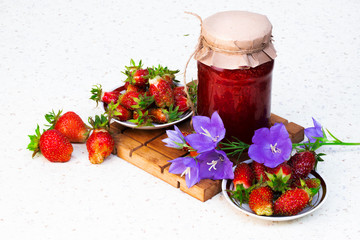 Strawberry berries on a white plate on a cutting Board with a jar of strawberry jam on a light background with bell flowers . Flatly. The view from the top.