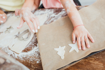 cropped view of child putting raw multi-shaped cookies on baking paper near mother and rolled dough, selective focus