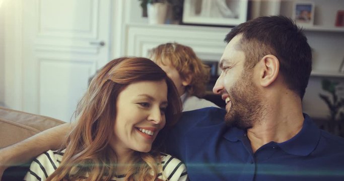 Young mom and dad are relaxing together sitting in living room on couch. Little son plays with parents hugging their necks.
