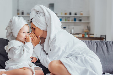 young woman telling secret to daughter while sitting together in bathrobes and towels on heads