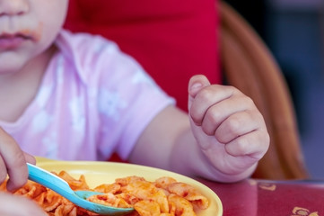 Toddler baby eating bowtie pasta with a plastic spoon, close-up. Cute baby hand.