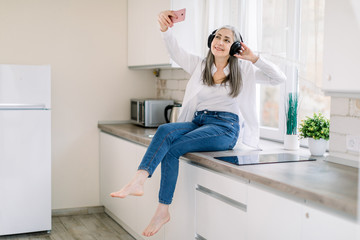 People at home concept. Happy Caucasian elderly woman with headphones holding mobile phone for making photo or video call, while sitting on kitchen countertop in modern kitchen