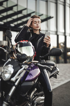 Beauty in a motorcycle jacket sits on a purple motorbike and looks at her phone
