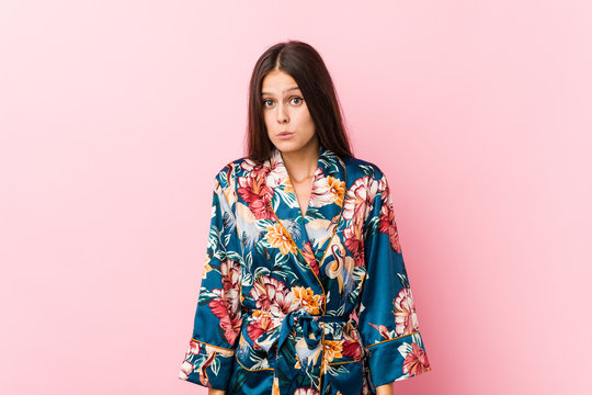 Young caucasian woman wearing a kimono pajama shrugs shoulders and open eyes confused.