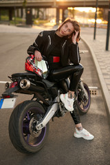 Naklejka na ściany i meble Young brunette girl in a black motorcycle jacket sits on a purple sport motorbike, with the sunset on the background