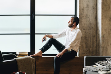 Mature man sitting barefoot on window sill, using smartphone and earphones