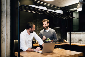 Mature friends working on laptop in kitchen, one eating salad