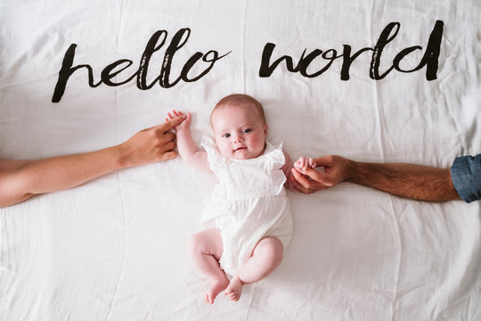 Happy cute baby girl holding hands of parents while lying on blanket under Hello World text at home