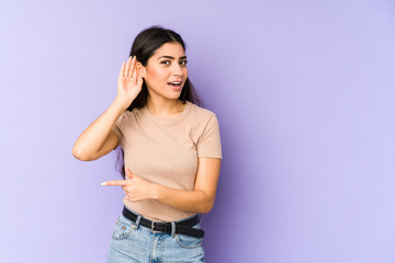 Young indian woman isolated on purple background trying to listening a gossip.