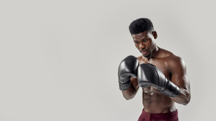 Young muscular african american male boxer looking down, wearing boxing gloves, standing isolated over grey background. Sports, workout, bodybuilding concept