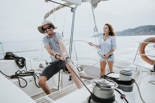 Man maneuvering with winch while woman driving sailboat against sky