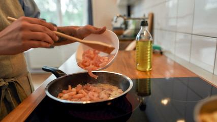 Close up of man adding shrimp to sauteed garlic and onion in the frying pan. Cook preparing dish with seafood. Mediterranean cuisine concept