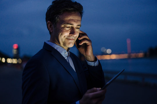 Mature businessman using tablet and smartphone at the riverbank at dusk