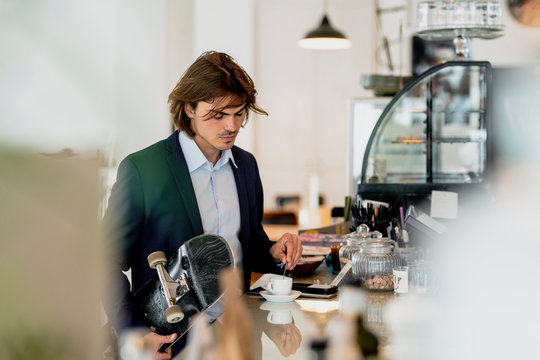 Businessman holding skateboard while mixing coffee on counter in cafe - Powered by Adobe