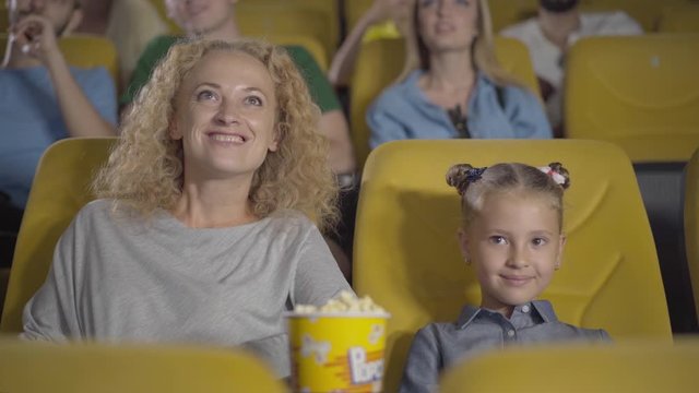 Embarrassed Caucasian mother closing daughter's eyes in cinema. Portrait of relaxed parent and child watching inappropriate film in movie theatre. Leisure, parenthood and childhood.