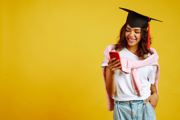 Graduate woman in a graduation hat posing with phone on a yellow background. African American woman...