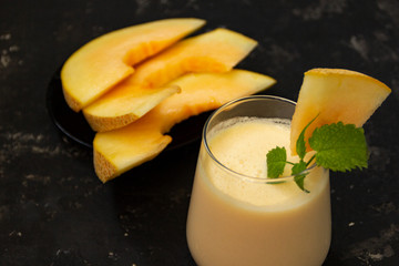 Melon milk shake on a black background. Ripe sliced melon is lying nearby. The smoothie glass is...