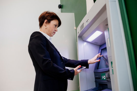 Businesswoman holding smart phone using ATM while standing by wall