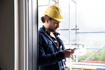 Construction worker wearing helmet using smart phone by window at constructing house