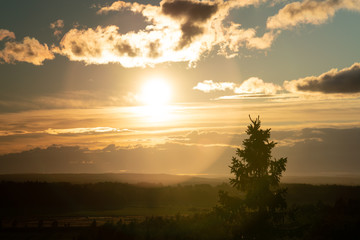 Aerial sunset photo above dark green northern scandinavian forest - round lens flare, clouds, fir tree top, warm foggy evening, yellow red colored sun light, Scandinavian mountains