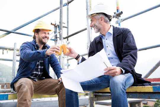 Architect and construction worker toasting drinks while sitting against clear sky at construction site