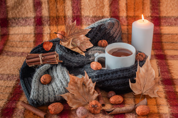 lit candle on old checkered blanket with orange red and brown patterns.dry leafs. autumn background.  dry fruits. cozy  woolly gray scarf. cup of coffee