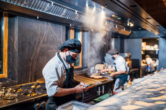 Chef wearing protective face mask using smartphone in restaurant kitchen