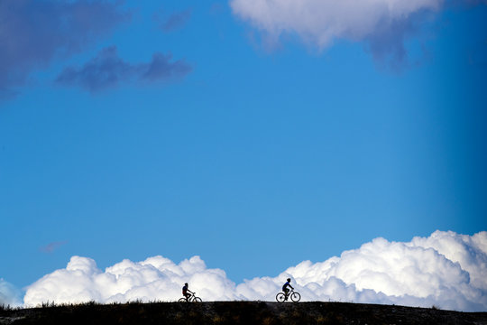 Silhouette cyclists riding bicycles on field against cloudy blue sky