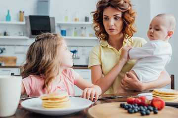 selective focus of mother holding son while looking at daughter sitting near plate with delicious pancakes