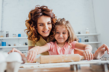 selective focus of child rolling out dough on table near young curly mother