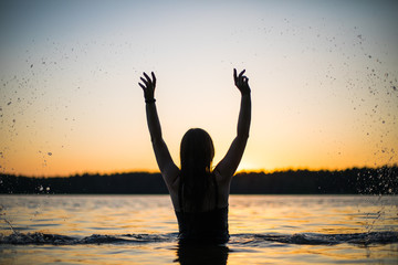 Beautiful Russian girl in a long black swimsuit swims outside the city on the lake in the rays of sunset or dawn. Splashing water on the background of the sun. Happy girl.