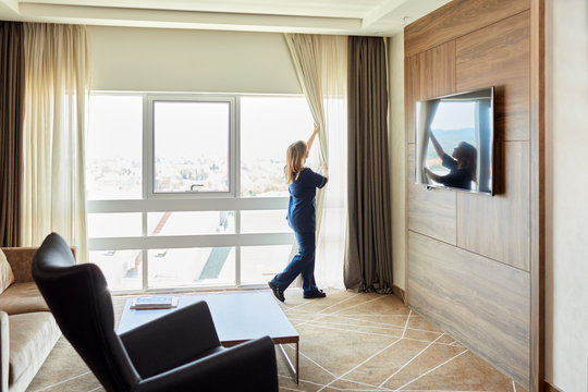 Chambermaid opening curtains of window in hotel bedroom
