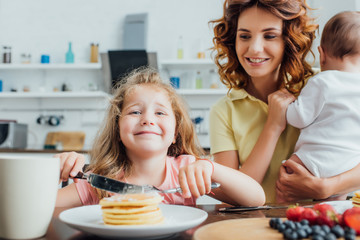 selective focus of girl looking at camera while eating pancakes near mother with infant