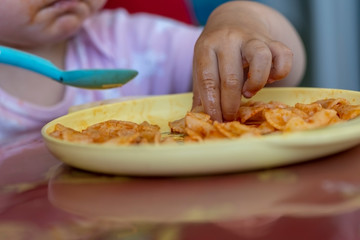 Toddler girl eating farfalle pasta with her fingers and a plastic spoon, close-up