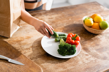 partial view of young woman holding cucumber near plate with broccoli and red bell pepper
