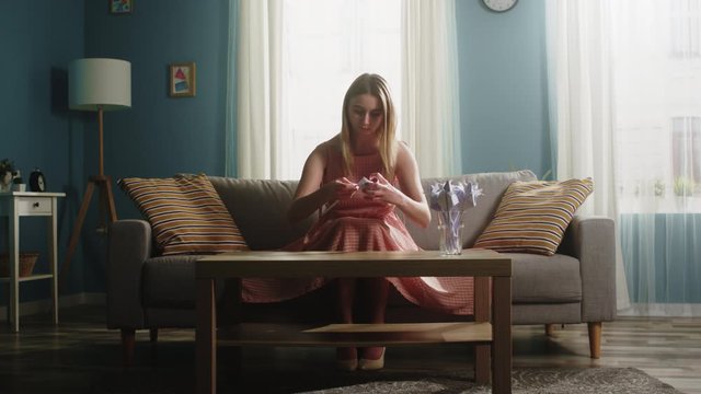 Young girl girl in a pink dress sits on the couch and folds origami from a white sheet of paper on a coffee table