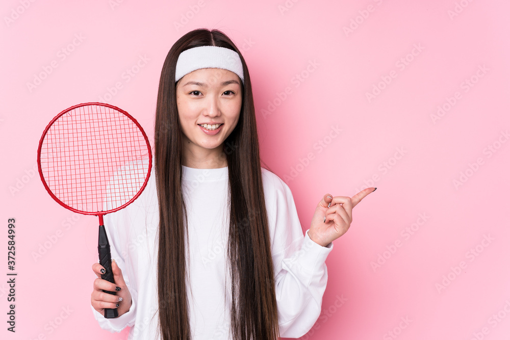 Wall mural young chinese woman playing badminton isolated smiling and pointing aside, showing something at blan