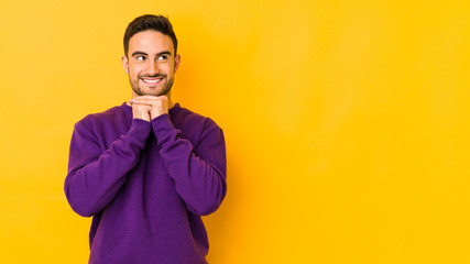 Young caucasian man isolated on yellow bakground keeps hands under chin, is looking happily aside.