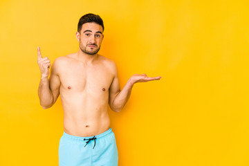 Young caucasian man with swimsuit isolated on yellow background Young caucasian man with trholding and showing a product on hand.