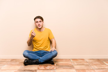 Young caucasian man sitting on the floor isolated pointing with finger at you as if inviting come closer.